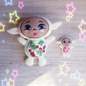 May include: Two plush toys shaped like sheep with embroidered faces. The larger sheep is white with a strawberry print and brown eyes. The smaller sheep is white with pink accents and black eyes.