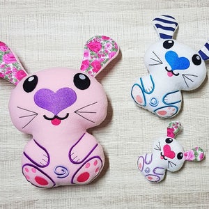 May include: Three plush bunny toys. The largest bunny is pink with floral print ears, purple heart nose, and pink feet. The middle bunny is white with blue and white striped ears, a blue heart nose, and blue feet. The smallest bunny is white with pink floral print ears, a pink heart nose, and purple feet.