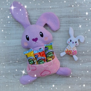 May include: A plush purple bunny rabbit holding a variety of colourful candy bars in a pink pouch. The bunny has a white heart on its nose and is sitting on a light wood background.