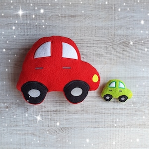 Puede incluir: Dos coches de peluche, uno rojo y otro verde. El coche rojo es más grande y tiene un detalle amarillo en el lateral. El coche verde es más pequeño y tiene un diseño sencillo.
