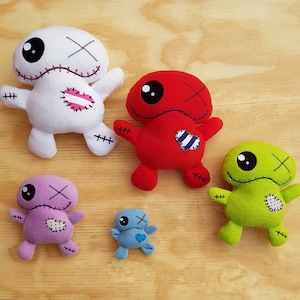 Puede incluir: Cinco juguetes de peluche hechos a mano en diferentes colores y diseños. Los juguetes tienen forma de personas con ojos, bocas y otros detalles cosidos. Los juguetes están todos de pie sobre una superficie de madera.