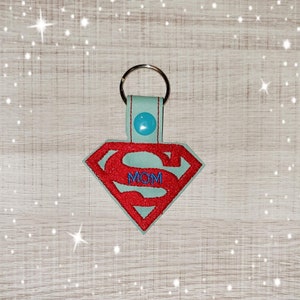 Könnte beinhalten: Ein hellblauer Schlüsselanhänger mit einem roten Superman-Logo und dem Wort "MOM" darin. Der Schlüsselanhänger hat einen silbernen Ring und einen blauen Knopf.