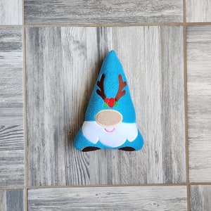 May include: A turquoise gnome-shaped plush doll with brown antlers and a red berry cluster. The gnome has a tan nose, white beard, and brown feet. The doll is a festive holiday decoration.
