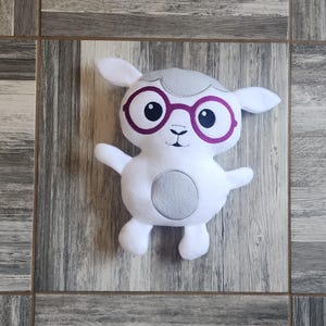 Puede incluir: Peluche de oveja blanca con detalles grises, que lleva gafas de montura rosa. La oveja tiene una sonrisa cosida y una mancha gris en el vientre.
