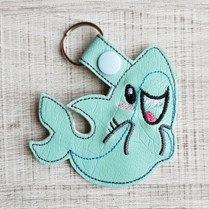 Könnte beinhalten: Ein hellblauer Haifisch-Schlüsselanhänger mit gestickten Details. Der Hai hat ein lächelndes Gesicht mit weißen Augen und einem rosa Mund. Der Schlüsselanhänger hat einen silbernen Ring und einen weißen Knopf.