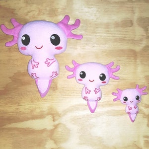 May include: Three pink plush axolotl toys of different sizes. The largest axolotl is about 10 inches tall, the middle axolotl is about 6 inches tall, and the smallest axolotl is about 4 inches tall. All three axolotls have pink and white eyes, a pink smile, and pink fins.