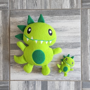 May include: Two green plush toy monsters with embroidered eyes and teeth. The larger monster has a green circle on its belly. The smaller monster is sitting on a wood-grain surface.