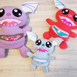 May include: Three plush toys with zippered pockets. The largest toy is purple with white wings and black eyes. The middle toy is red with blue wings and black eyes. The smallest toy is blue with white wings and black eyes.