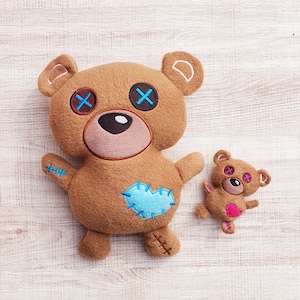 May include: Two brown plush teddy bears with embroidered details. The larger bear has blue X's for eyes and a blue heart patch. The smaller bear has pink X's for eyes and a pink heart patch.