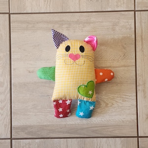 May include: A yellow and white checkered cat plush toy with colorful fabric accents. The cat has a pink heart nose, black eyes, and a green heart on its chest.