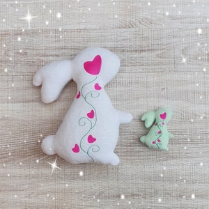 May include: Two plush bunny rabbits. The larger one is white with a pink heart and vine design. The smaller bunny is light green with a similar heart design. Both are on a light wood-grain surface.