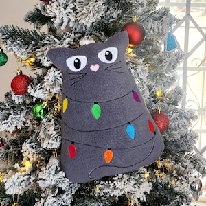 May include: A grey cat-shaped plush toy with embroidered eyes, whiskers, and a pink heart nose. The cat is decorated with colourful string lights, resembling a Christmas tree.