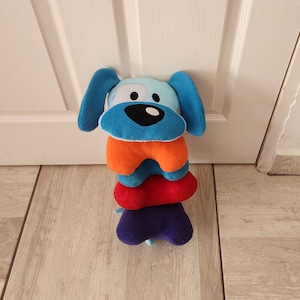 May include: A blue, orange, red, and purple plush dog toy with a smiling face. The toy is made of soft fabric and is designed for children.