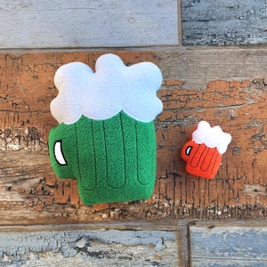 Può includere: Due giocattoli di peluche a forma di boccali di birra. Il boccale più grande è verde con la schiuma bianca in cima. Il boccale più piccolo è rosso con la schiuma bianca in cima.
