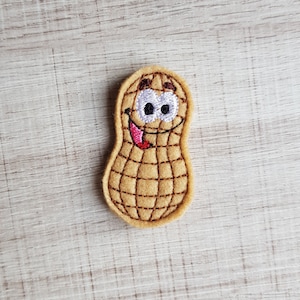 Puede incluir: Un cacahuete de fieltro marrón con una cara sonriente, ojos blancos y una lengua rosa que sobresale. El cacahuete tiene una cáscara de fieltro marrón con un patrón de cuadrícula cosido.