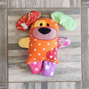 May include: A colorful patchwork dog toy with orange, green, yellow, pink, and white fabric. The dog has a black nose and eyes, and a red heart on its chest.