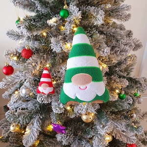 May include: Two plush gnomes hanging on a flocked Christmas tree. The larger gnome is green and white with a white beard and a pink nose. The smaller gnome is red and white with a white beard and a pink nose.