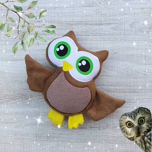 May include: A brown plush owl with large green eyes and a yellow beak. The owl has its wings spread out and is standing on a white wooden surface.