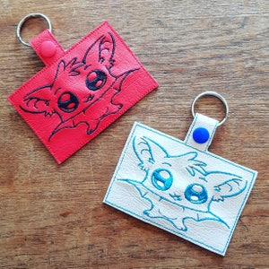 May include: Two keychains with a cute cartoon bat design. One keychain is red with a black bat and the other is white with a blue bat.