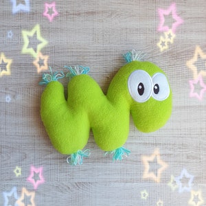 May include: A lime green plush toy shaped like a stylized worm with large white-rimmed eyes and blue and white tufts. The toy is on a light wood-grain surface with pastel star accents.