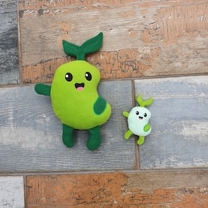 May include: Two plush toys shaped like green beans with smiling faces. The larger bean has a green stem and leaves. The smaller bean has a green stem and a single leaf. Both beans have white eyes and a pink mouth.