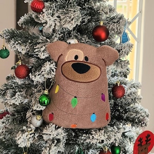 May include: A brown plush dog ornament with a stitched-on face and a string of colourful lights around its neck. The ornament is hanging on a flocked Christmas tree.