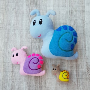 Pode incluir: Três caracóis de peluche, um azul, um rosa e um castanho. O caracol azul tem uma concha azul com um desenho de espiral branco. O caracol rosa tem uma concha roxa com um desenho de espiral rosa. O caracol castanho tem uma concha amarela com um desenho de espiral branco.