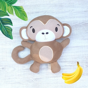 Könnte beinhalten: Ein braunes Plüsch-Affenspielzeug mit weißem Gesicht und schwarzen Augen. Der Affe hat einen langen Schwanz und sitzt auf einer weißen Holzoberfläche. Neben dem Affen liegen drei gelbe Bananen.