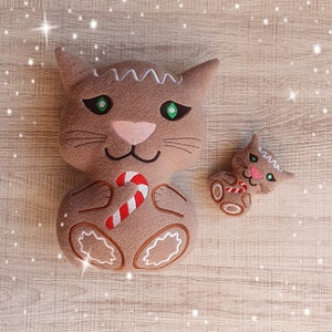 Peut inclure: Deux jouets en peluche en forme de chat en pain d'épices. Le plus grand chat tient une canne en sucre. Les deux chats ont des yeux verts et une fourrure brune.