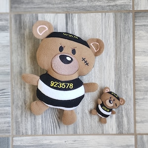 Puede incluir: Dos osos de peluche marrones que llevan trajes de prisioneros a rayas blancas y negras. El oso más grande lleva una diadema negra con el texto "923578" y el oso más pequeño lleva el texto "923578" en el pecho.