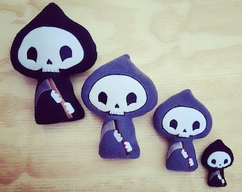 Puede incluir: Cuatro peluches con forma de Parca en diferentes tamaños. La más grande es negra con una calavera blanca y una guadaña marrón. Las otras tres son moradas con calaveras blancas y guadañas marrones.