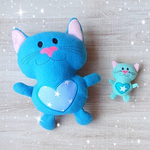 Puede incluir: Dos juguetes de peluche de gato azul con corazones y estrellas bordadas. El gato más grande tiene orejas rosas y un corazón blanco con estrellas azules. El gato más pequeño tiene orejas verdes y una estrella blanca en el centro de su corazón.
