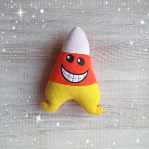 Könnte beinhalten: Ein Plüschtier in Form eines Zuckermais mit einem lächelnden Gesicht. Das Spielzeug ist orange, gelb und weiß.