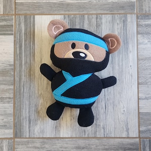 May include: A plush toy bear dressed as a ninja. The bear is wearing a black suit with a blue sash and a blue headband. The bear has brown ears and a brown nose.