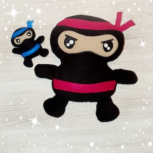 以下が含まれることがあります： ピンクとブルーのアクセントが付いた、黒いフェルト製の忍者フィギュア2体。大きな忍者はピンクのヘッドバンドを付けており、真剣な表情をしています。小さな忍者は青いヘッドバンドを付けており、より遊び心のある表情をしています。