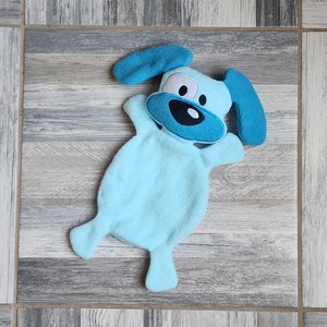 May include: A light blue plush dog toy with a blue embroidered face and ears. The toy is lying on a wood-grain surface.