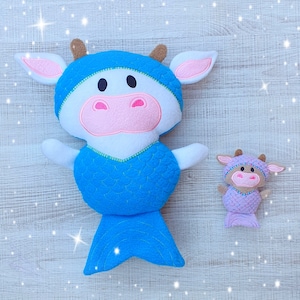 May include: Two plush cow toys. The larger cow is white with a blue mermaid tail and blue accents. The smaller cow is lavender with a mermaid tail. Both have pink noses and ears, brown horns, and black eyes. The toys are on a light wood-grain background.