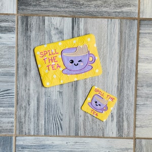 Könnte beinhalten: Zwei gelbe Untersetzer mit weißem Punktmuster und dem Text "Spill the Tea" in Pink. Jeder Untersetzer zeigt eine lilafarbene Cartoon-Teetasse mit einem lächelnden Gesicht.