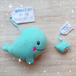 May include: Two plush whale toys with a white tag that says "Whale you be my Valentine". The larger whale is light blue and the smaller whale is a darker blue.
