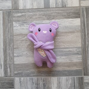May include: A purple plush toy bear wearing a purple hooded blanket with a floral heart design. The bear has a cute embroidered face with pink cheeks.