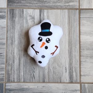 May include: A white plush snowman pillow with black eyes, a carrot nose, and a black top hat with a blue band. The snowman has two black twig arms.