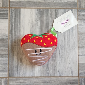May include: A plush heart-shaped strawberry with chocolate frosting and a smiling face. A tag attached to the strawberry reads "I love you berry much".