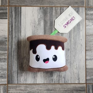 May include: A plush s'more pillow with a smiling face, brown chocolate, and white marshmallow. The pillow is on a woodgrain background with a tag that says "I love you s'more!"