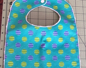 May include: A turquoise blue baby bib with a white trim. The bib has a repeating pattern of green and purple striped circles.