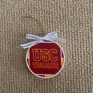 May include: A red and yellow round ornament with the text "USC Trojans" in yellow lettering. The ornament has a white ribbon attached to it.