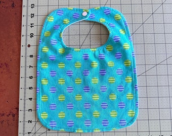 May include: A turquoise blue baby bib with a white snap closure. The bib has a pattern of green and purple stripes inside of blue circles.
