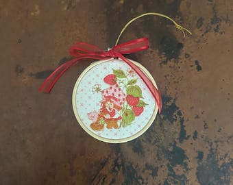 Könnte beinhalten: Runder, bestickter Schmuck mit einer Zeichentrickfigur, Erdbeeren und einer Katze. Das Design ist in einem Holzreifen gefasst, mit einem roten Band und einer goldenen Schnur zum Aufhängen. Der Hintergrund ist ein marmoriertes Braun.