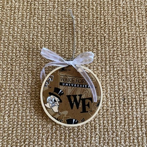 May include: A round, fabric-covered ornament featuring the Wake Forest University logo and text. The ornament has a cream-colored wooden frame, a white ribbon, and a silver hanging loop. The background fabric is brown with the university's logo.