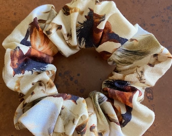 May include: A beige scrunchie featuring a horse print in brown, black, and tan hues. The fabric is gathered and elasticated, giving a ruffled look. This hair accessory is designed to secure hair in a ponytail or bun.