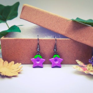 May include: A pair of purple flower earrings with green leaves hanging from silver hooks. The earrings are displayed in a brown box with a white interior.